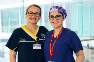 Two female nurses stand beside each other. The nurse on the right wears a surgical cap.
