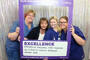 Four women stand inside a frame that reads 'South Metropolitan Health Service: Excellence'. A handwritten message on the board reads 'Operating theatres work together as a team to provide excellent care.'