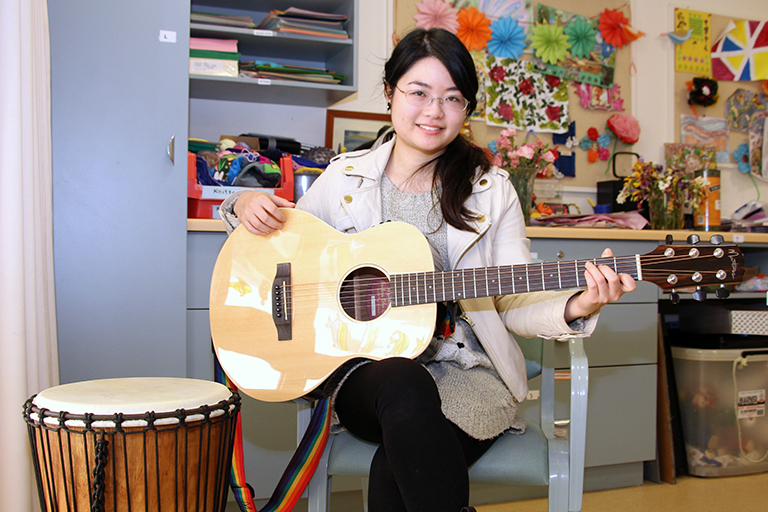 A young woman seated in a chair holds a guitar. A drum is next to the chair.
