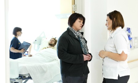 A nurse standing by the bedside of a male patient, with two women standing talking in a nearby doorway