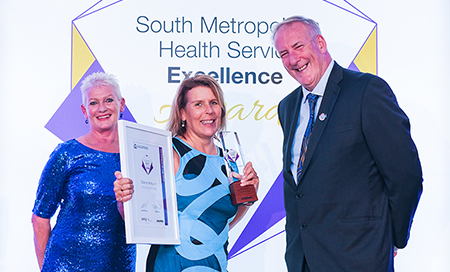 Two women and a man stand in front of a banner that reads South Metropolitan Health Service Awards. One woman holds a certificate and an award trophy. 