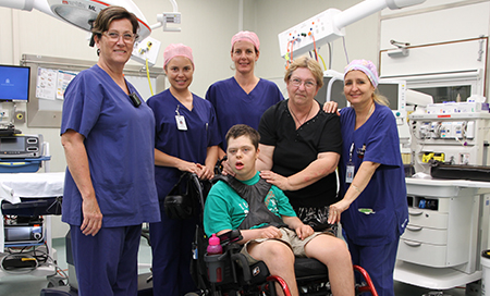 A young man in a wheelchair and a woman stand in a hospital theatre with four female theatre staff.