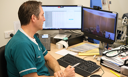A male health professional sits in front of computer and conducts an online conversation with two other people who are visible on the computer screen.
